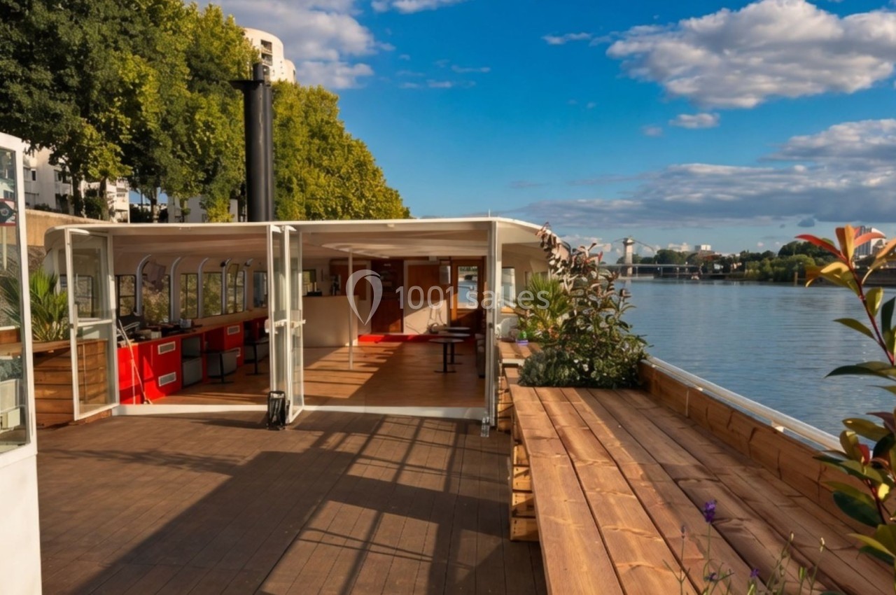 Terrasse en bois d'une péniche aménagée avec vue sur une rivière, entourée de végétation et sous un ciel dégagé.