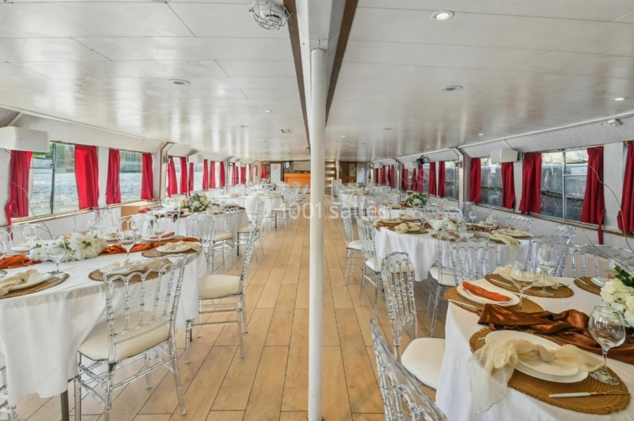 Salle de réception sur un bateau, décorée avec des tables rondes, nappes blanches, chaises transparentes et fleurs.