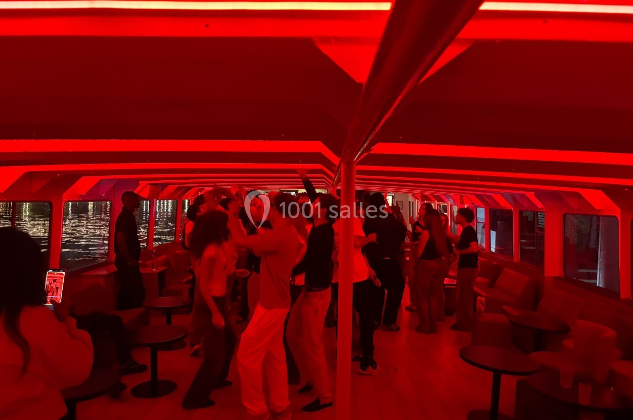 Des personnes dansent dans un espace intérieur éclairé par des lumières rouges, avec des tables et des banquettes autour.