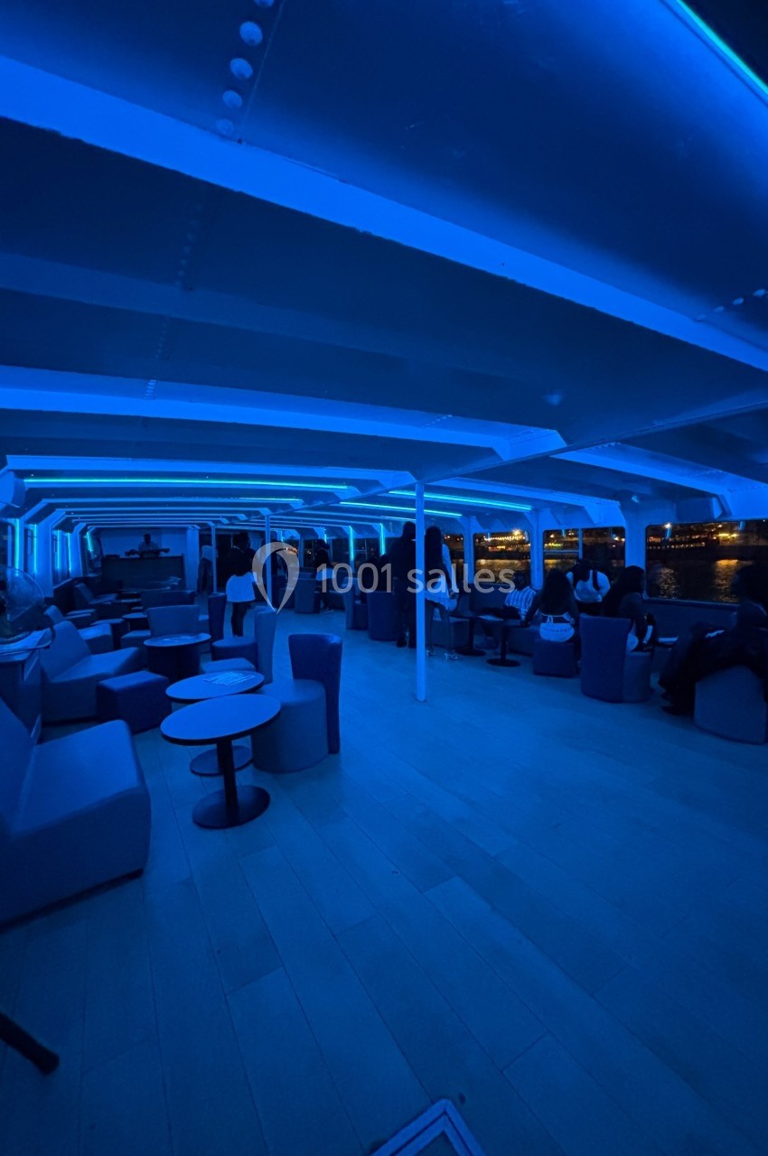 Intérieur d'un bateau avec éclairage bleu, des passagers assis près de tables et des fenêtres donnant sur l'eau.