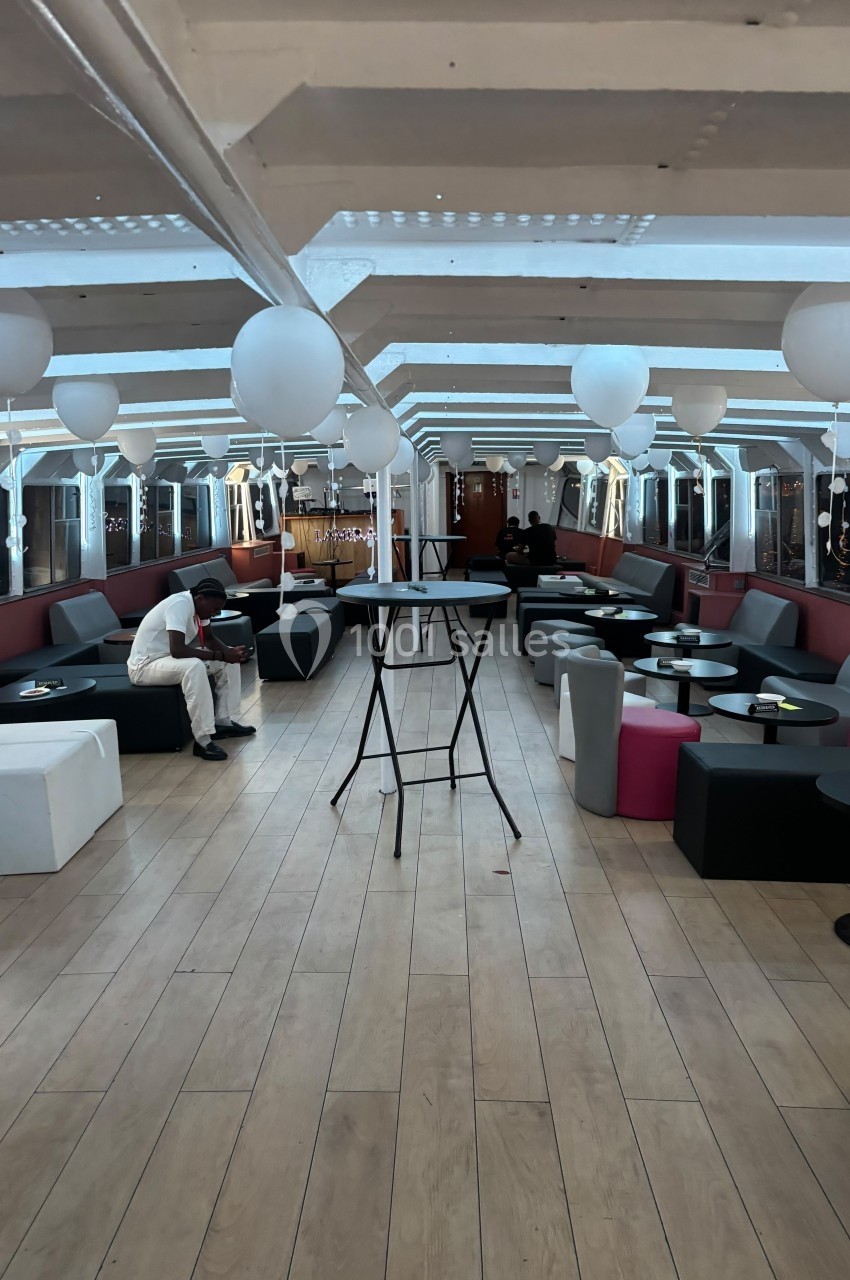 Salle lumineuse avec parquet, tables hautes et basses, canapés, ballons blancs suspendus et une personne assise.