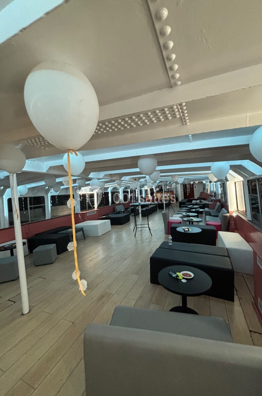 Salle de réception sur un bateau, décorée avec des ballons blancs, tables basses et banquettes modernes.