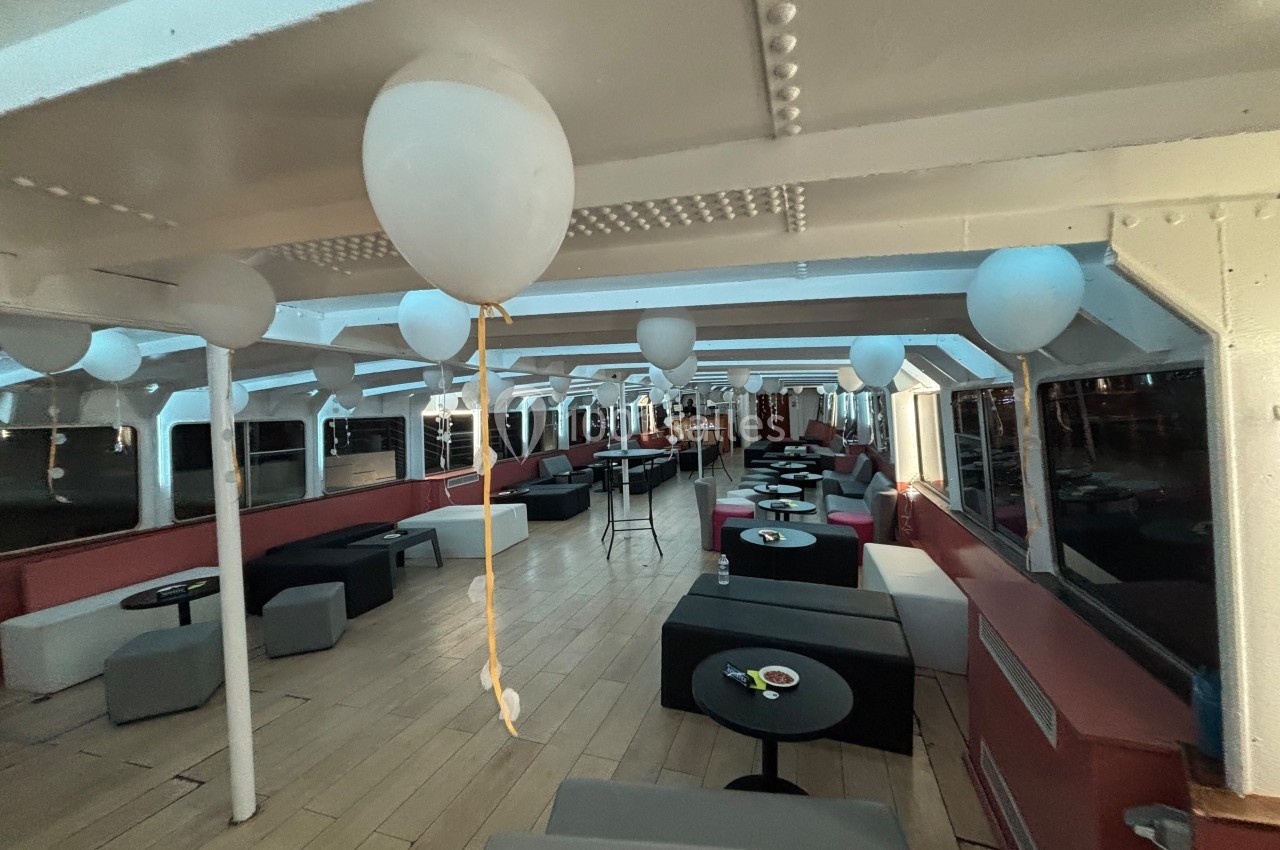 Salle intérieure d'un bateau aménagée avec des tables, des fauteuils et des ballons suspendus au plafond.