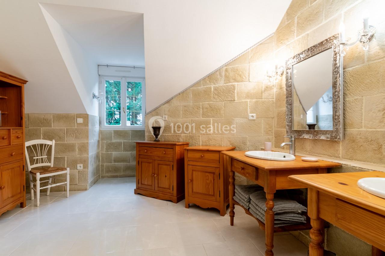 Salle de bain lumineuse avec meubles en bois, miroir encadré, murs en pierre beige et fenêtre donnant sur des arbres.