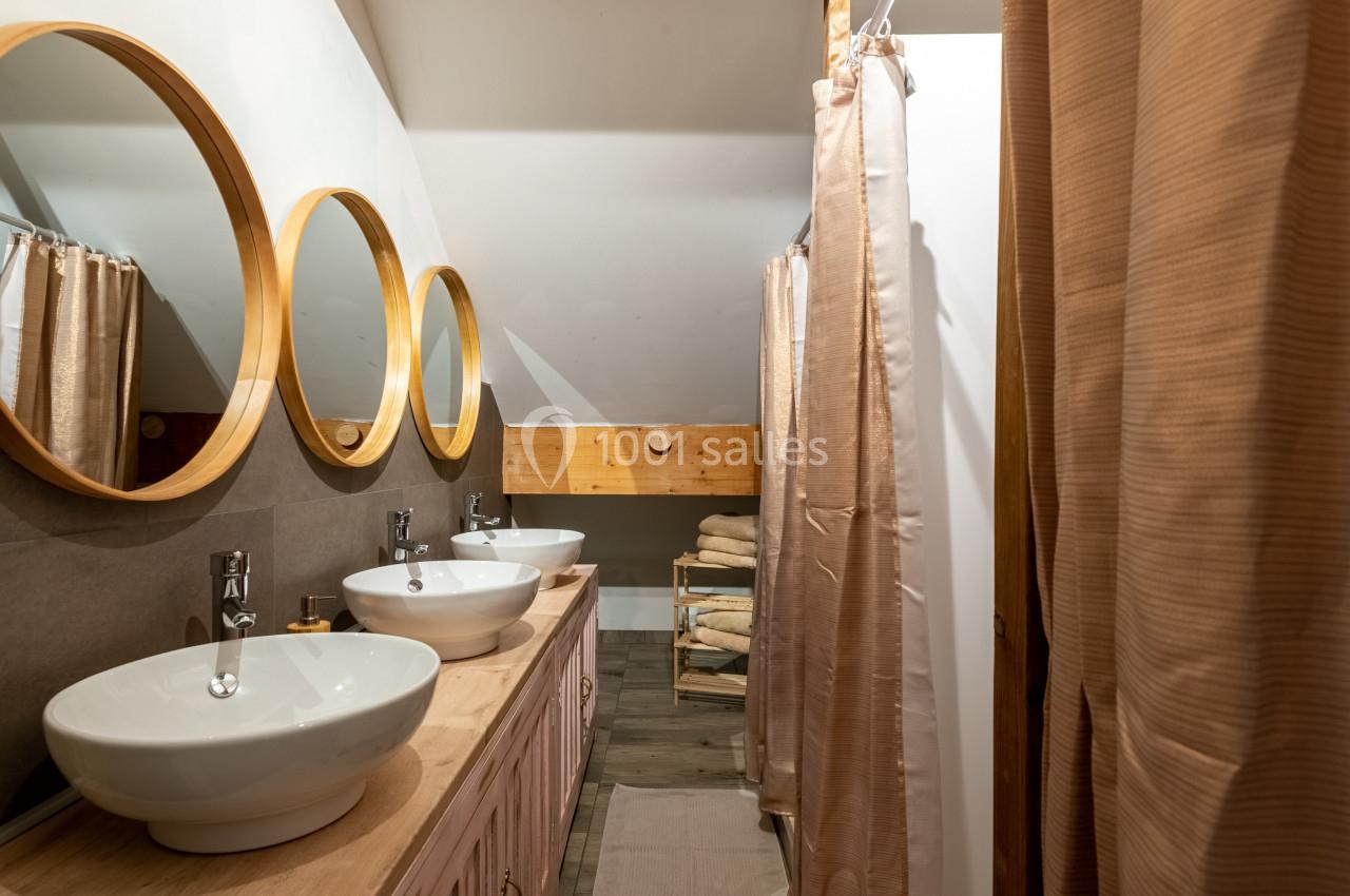 Salle de bain avec trois lavabos blancs, miroirs ronds en bois et rideaux beige séparant une douche.