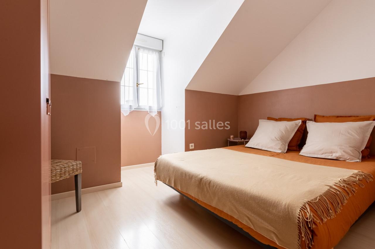 Chambre mansardée avec lit double, murs beige et marron, lumière naturelle entrant par une fenêtre avec rideaux blancs.