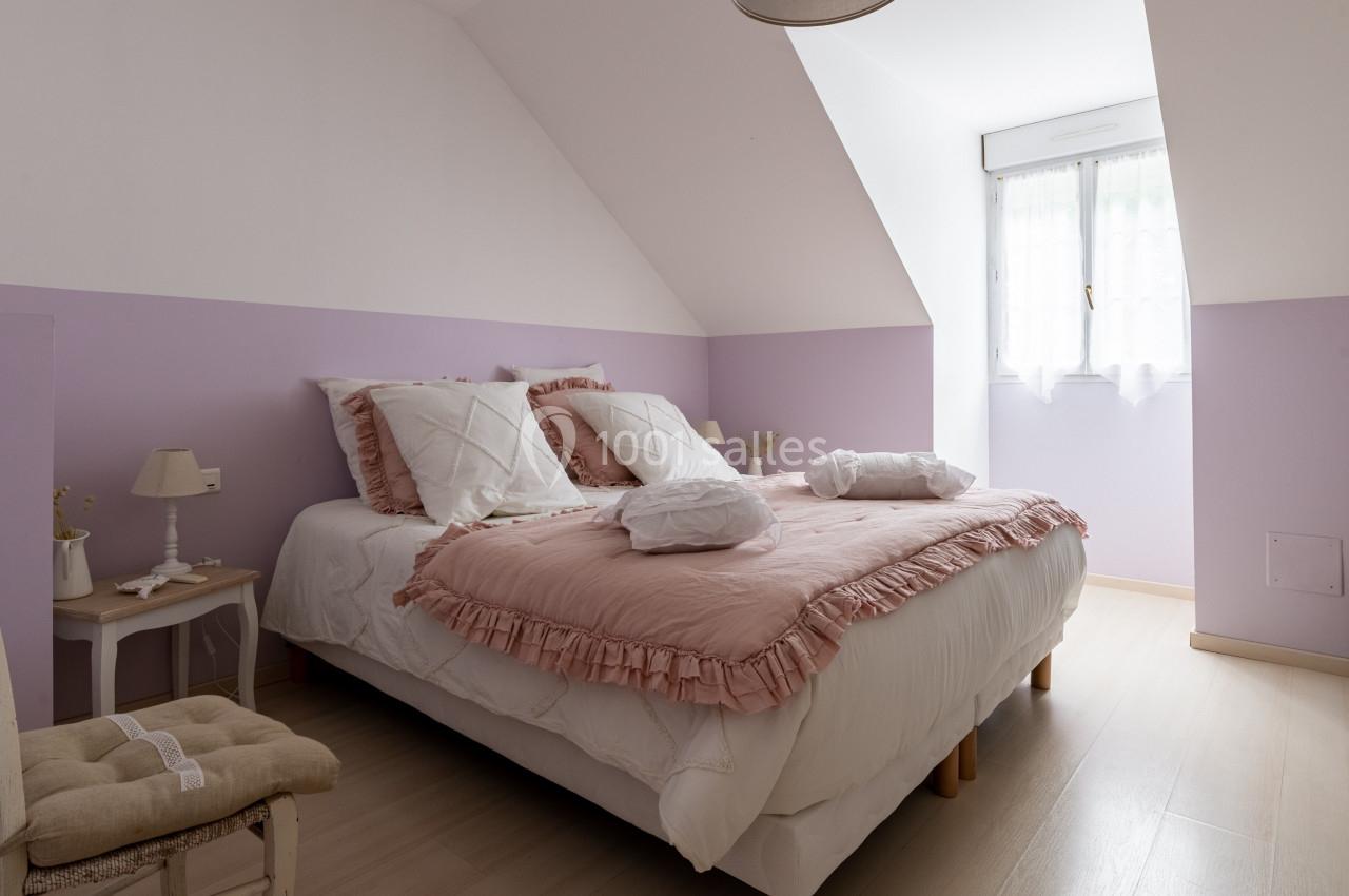 Chambre lumineuse avec lit double, draps roses et blancs, murs lilas, fenêtre avec rideaux légers et mobilier simple.