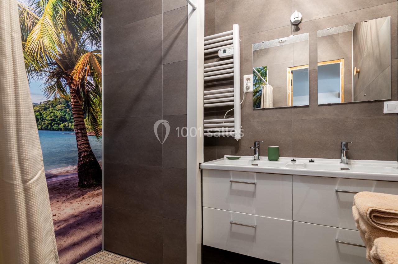 Salle de bain moderne avec lavabo double, miroir, radiateur sèche-serviettes et paroi décorée d'une plage tropicale.