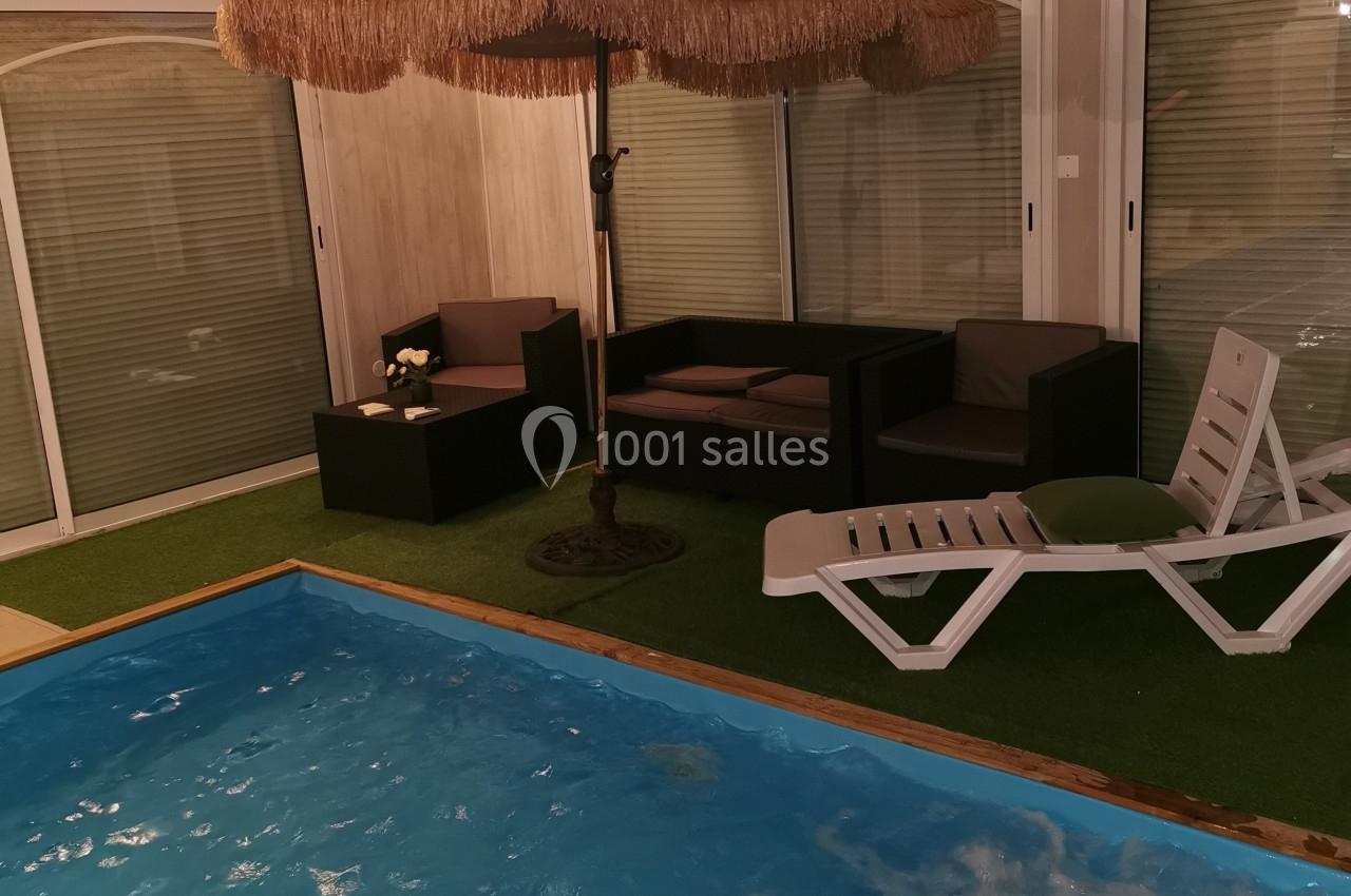 Espace intérieur avec piscine, parasol en paille, transat blanc et mobilier de jardin sur gazon artificiel.