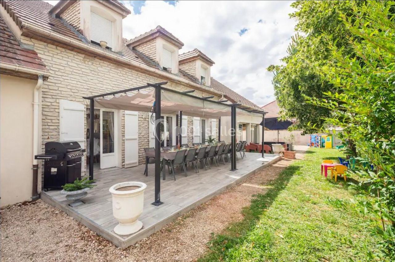Terrasse couverte avec table et chaises devant une maison en pierre, entourée de verdure et d'un espace de jeux pour enfants.