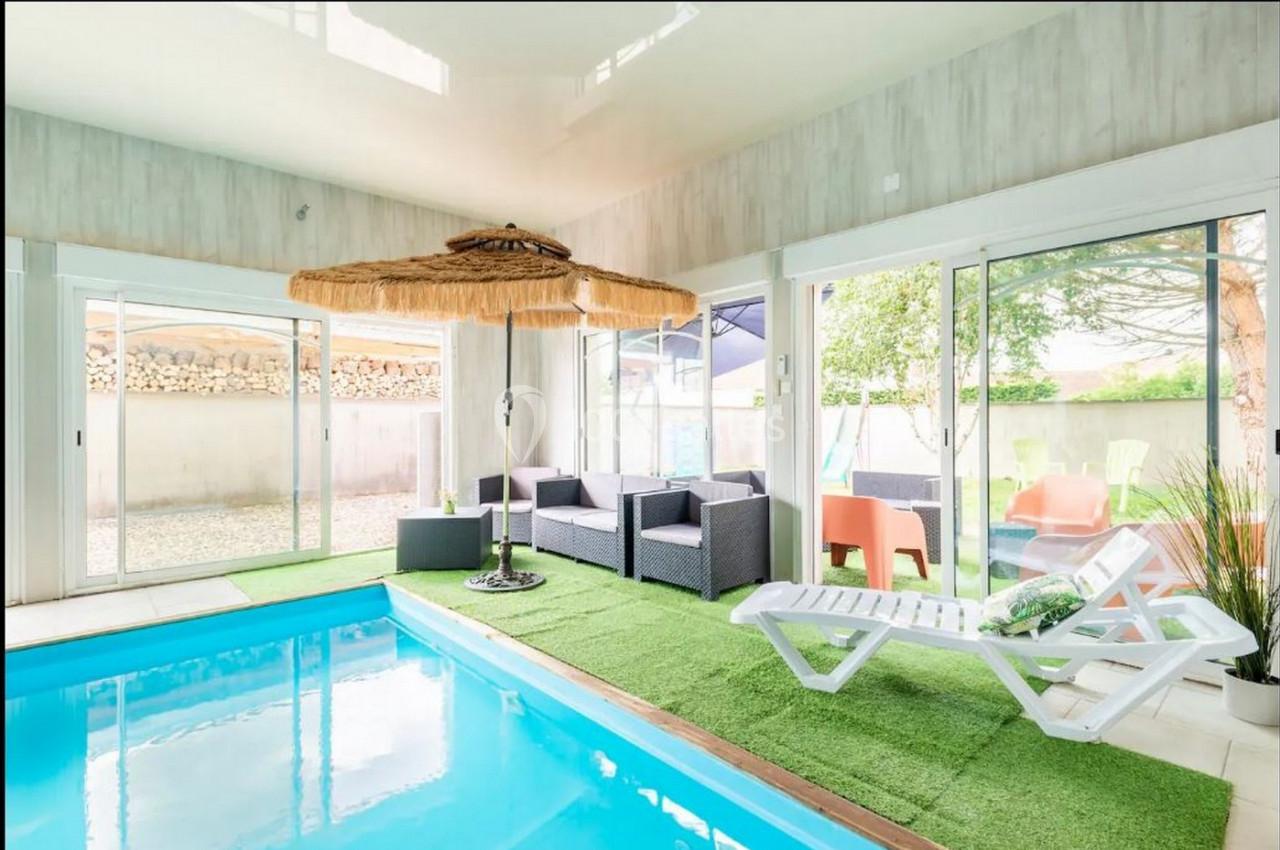 Piscine intérieure avec chaises longues, parasol en paille et mobilier de jardin sur gazon synthétique, vue sur l'extérieur.