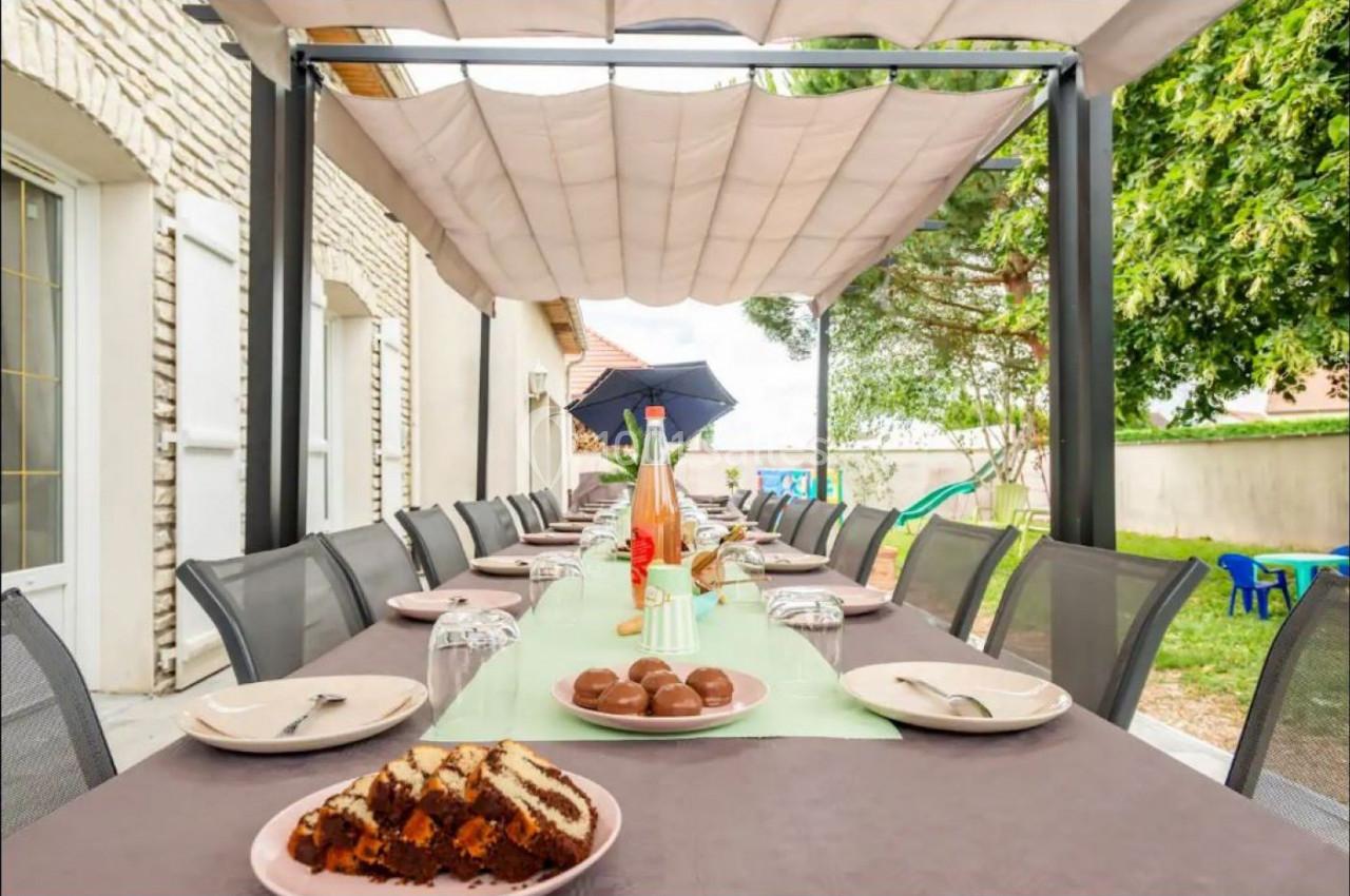 Grande table extérieure dressée avec vaisselle, desserts et boissons, sous une pergola dans un jardin arboré.