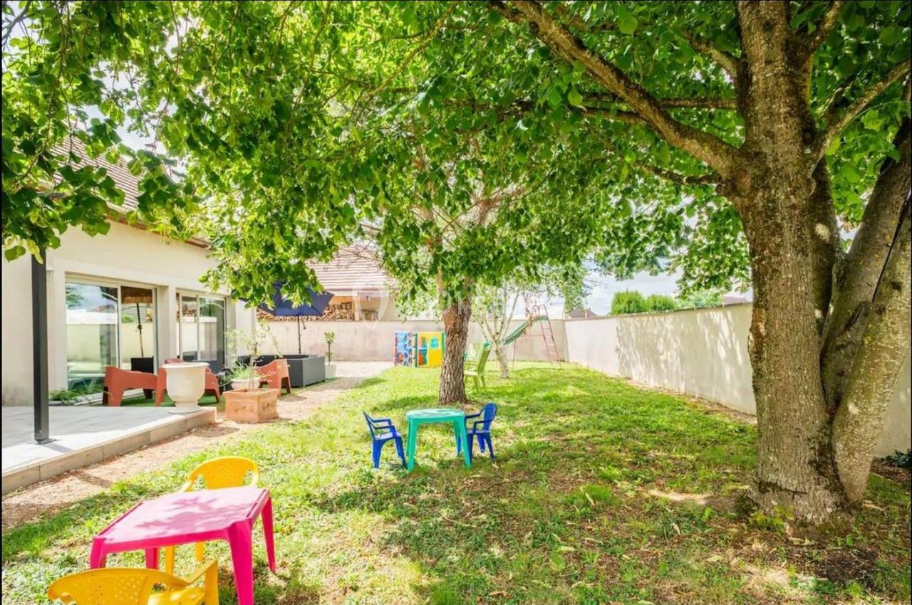 Jardin avec pelouse, arbres, tables et chaises colorées pour enfants, adjacent à une maison avec terrasse.
