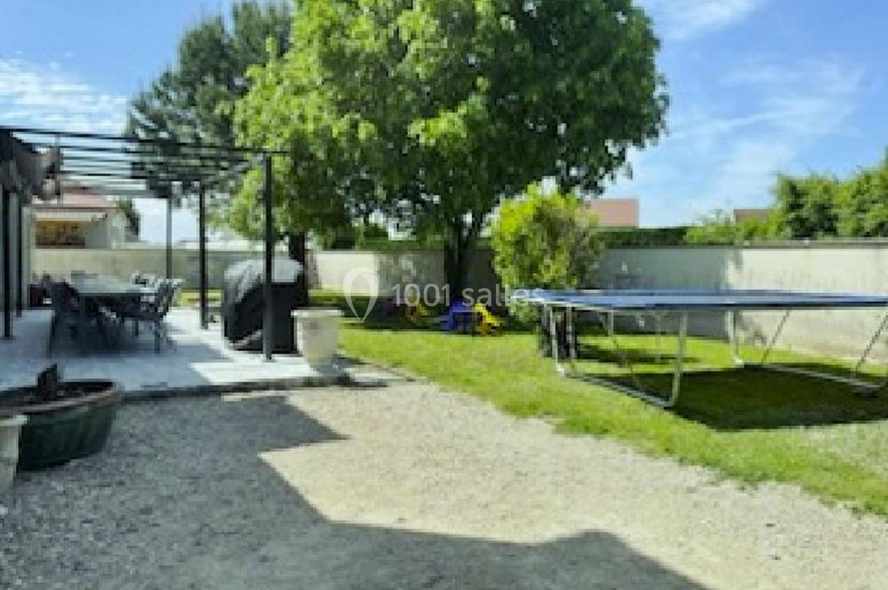 Jardin avec terrasse ombragée, trampoline, pelouse, arbre et clôture en arrière-plan sous un ciel dégagé.