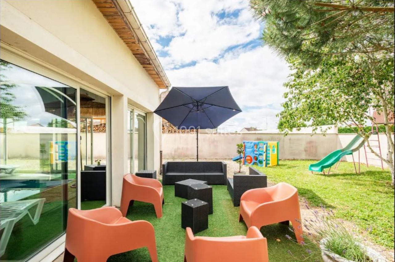 Mobilier de jardin avec fauteuils orange, table noire et parasol, installé sur une pelouse avec toboggan en arrière-plan.