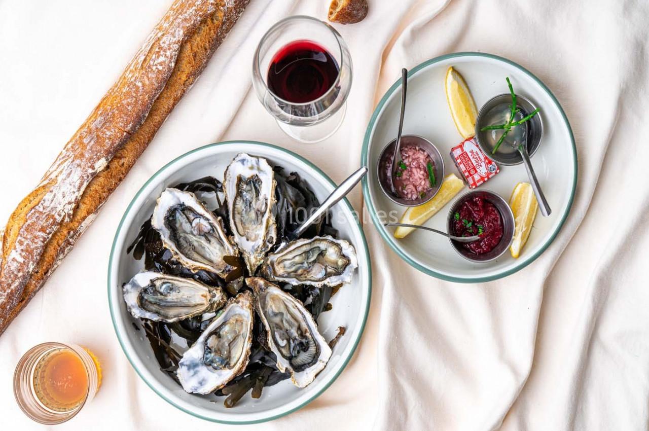 Assiette d'huîtres ouvertes avec accompagnements, verre de vin rouge, baguette et sauce disposés sur une nappe claire.