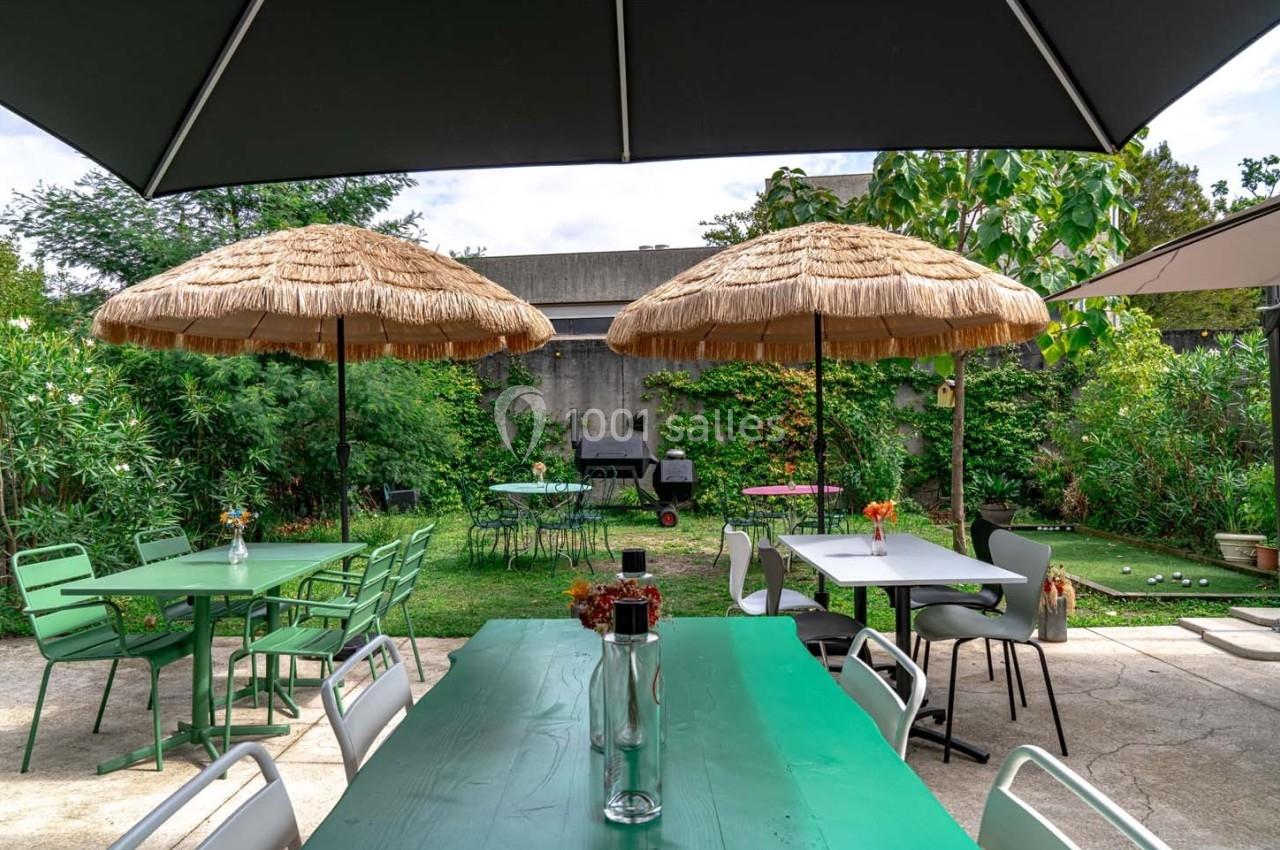 Terrasse extérieure avec tables, chaises, parasols en paille et jardin verdoyant en arrière-plan.