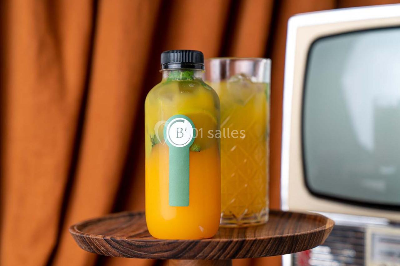 Bouteille de jus orange avec menthe posée sur un plateau en bois, devant un verre rempli et une télévision rétro.