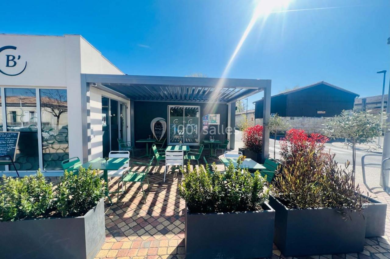 Terrasse extérieure d'un café avec tables et chaises vertes, entourée de jardinières sous un ciel ensoleillé.