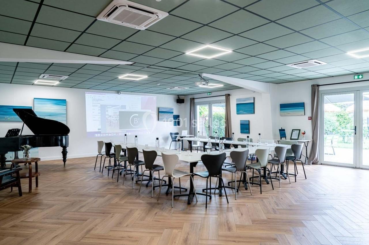 Salle de réunion lumineuse avec tables disposées en U, écran de projection, piano et vue sur un jardin par des baies vitrées.
