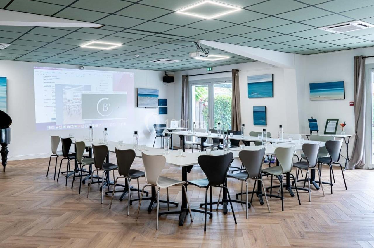 Salle de réunion lumineuse avec tables disposées en U, chaises modernes, écran de projection et décorations murales.