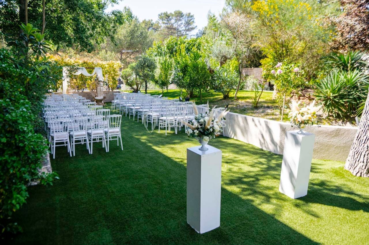 Allée centrale bordée de chaises blanches sur pelouse, menant à une arche décorée pour une cérémonie en extérieur.