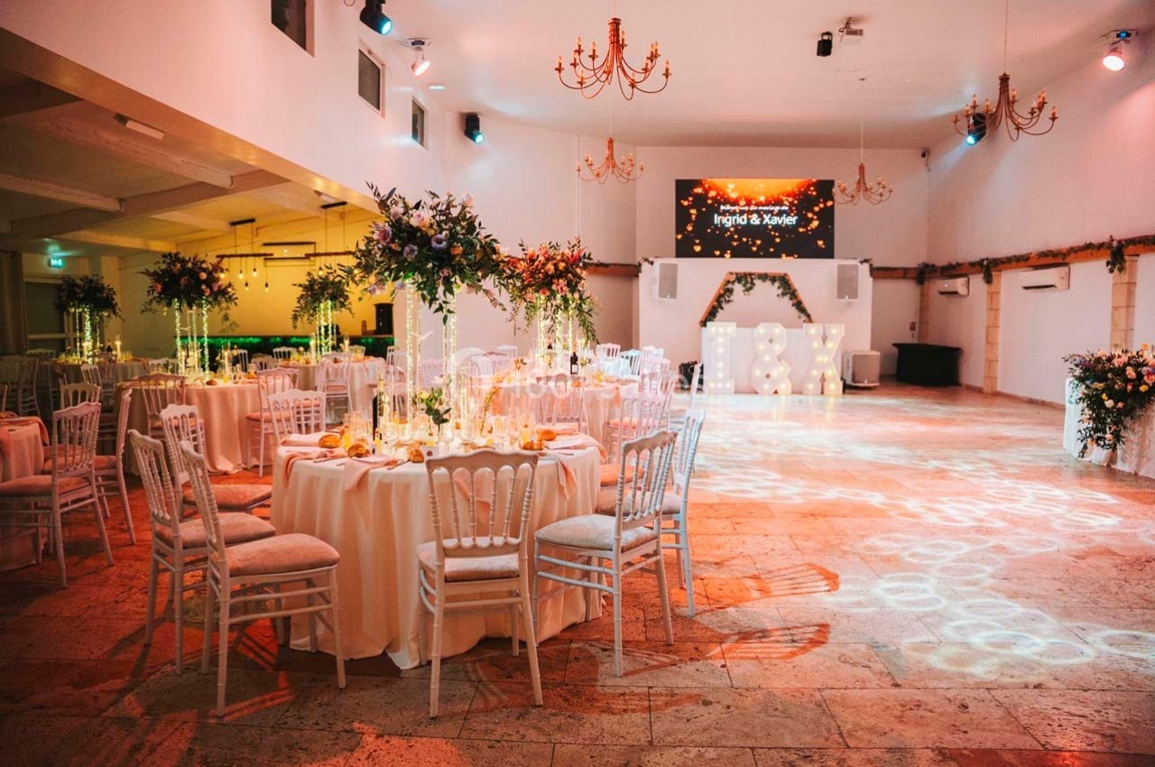 Salle de réception décorée avec des tables rondes, des chaises blanches et des arrangements floraux, éclairée par des…