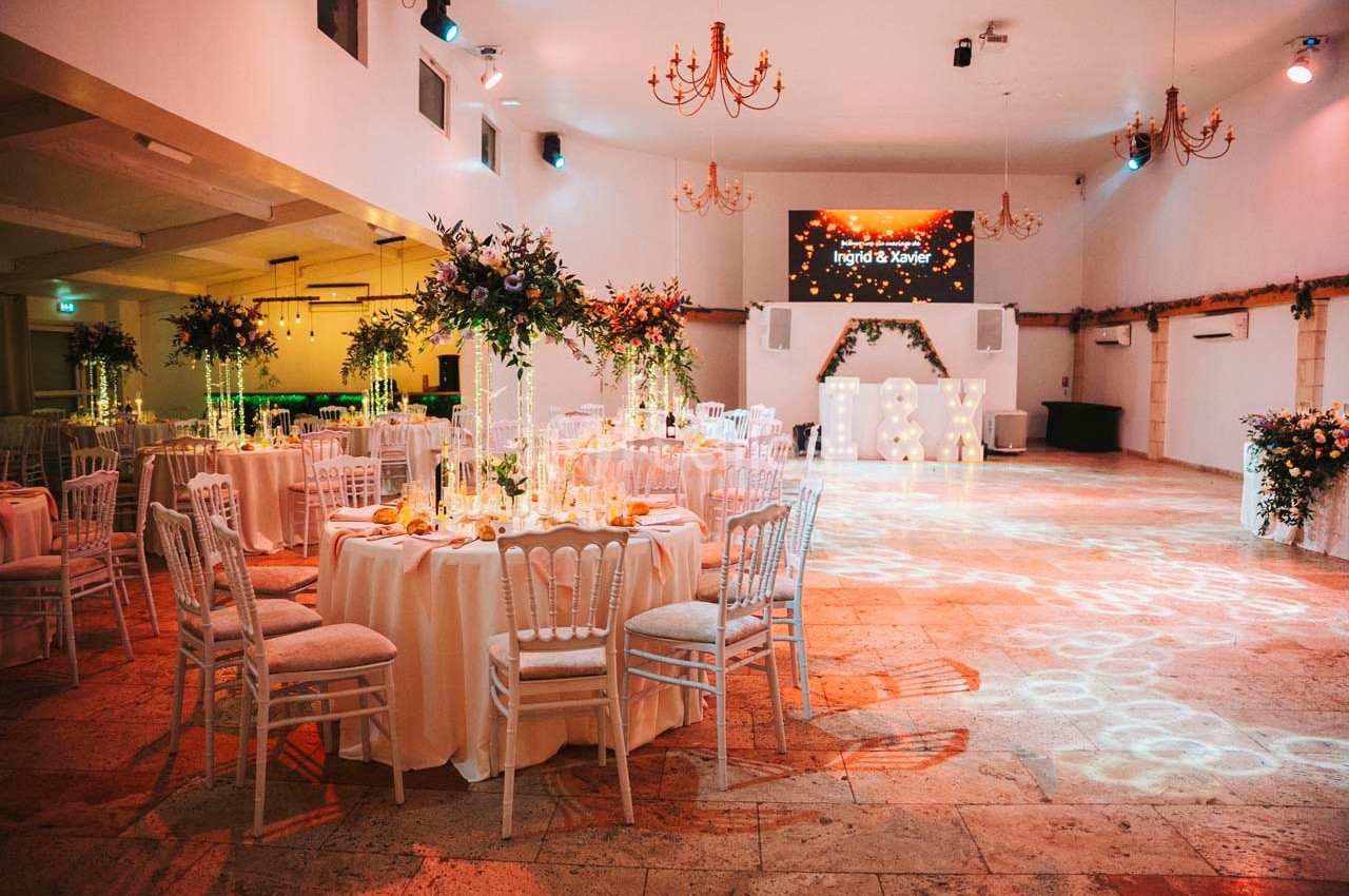 Salle de réception décorée avec des tables rondes, des chaises blanches et des arrangements floraux, éclairée par des…