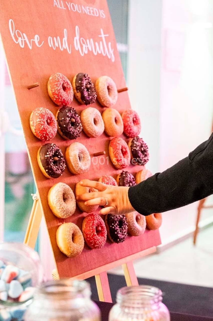 Mur en bois avec des donuts accrochés, une main en saisit un, pots de bonbons visibles au premier plan.