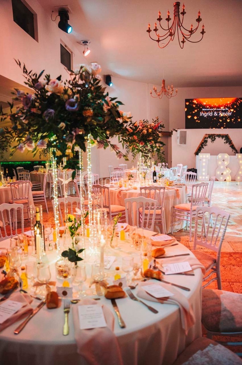 Salle de réception décorée avec des tables rondes, centres de table fleuris et éclairages chaleureux.