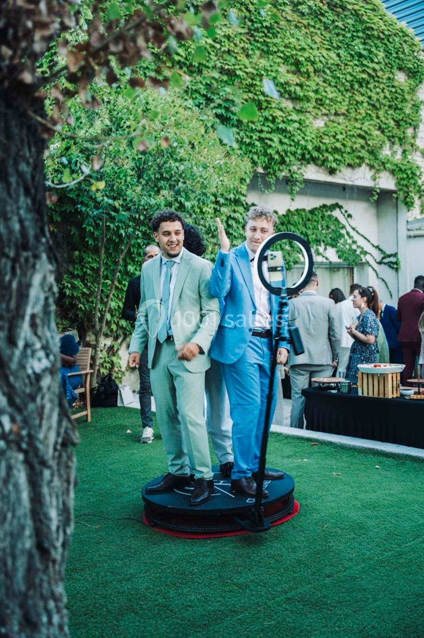 Deux hommes en costume posent sur une plateforme tournante dans un jardin, entourés de verdure et de convives.