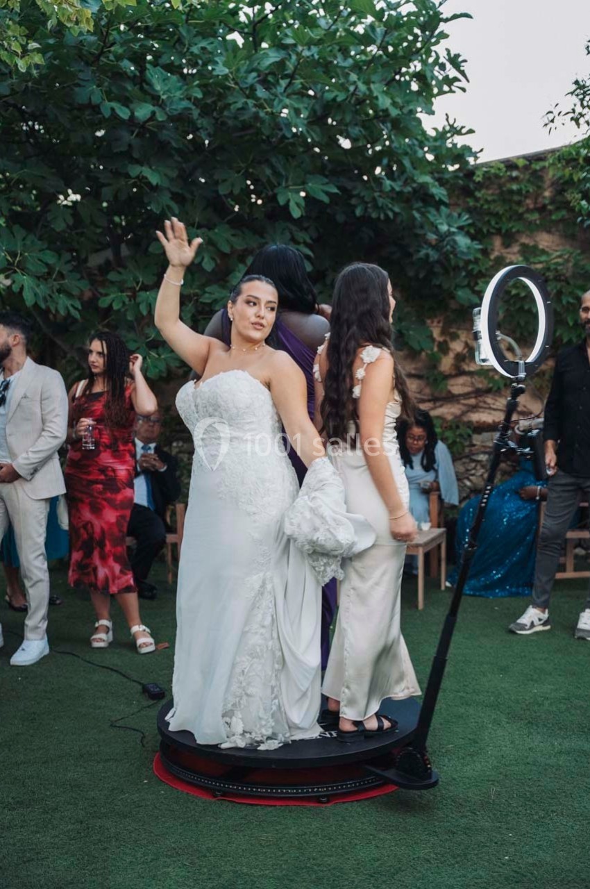 Une mariée en robe blanche pose sur une plateforme tournante lors d'un événement en extérieur, entourée d'invités.