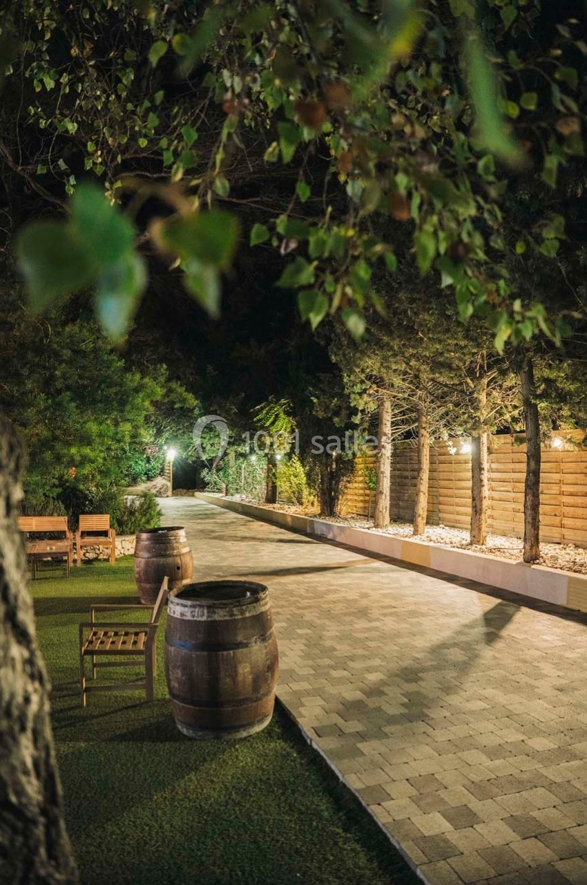 Allée pavée bordée d'arbres et de lumières, avec des tonneaux et des bancs en bois sur un espace gazonné.