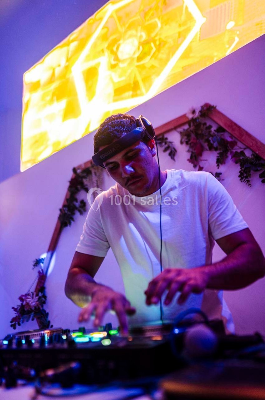 Un DJ en t-shirt blanc mixe de la musique sur une table de mixage, sous un éclairage coloré et un écran lumineux.
