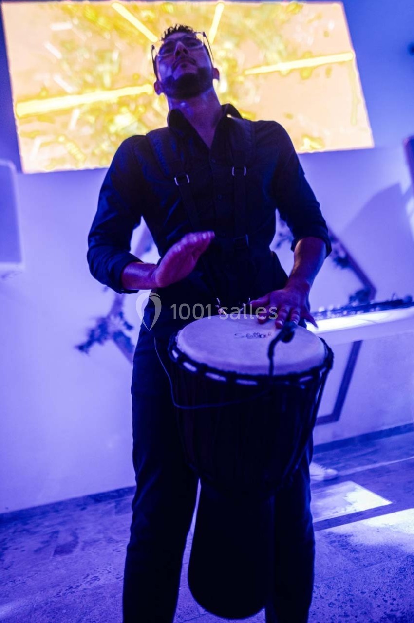 Un homme joue du djembé dans une ambiance lumineuse bleutée, devant un écran coloré en arrière-plan.