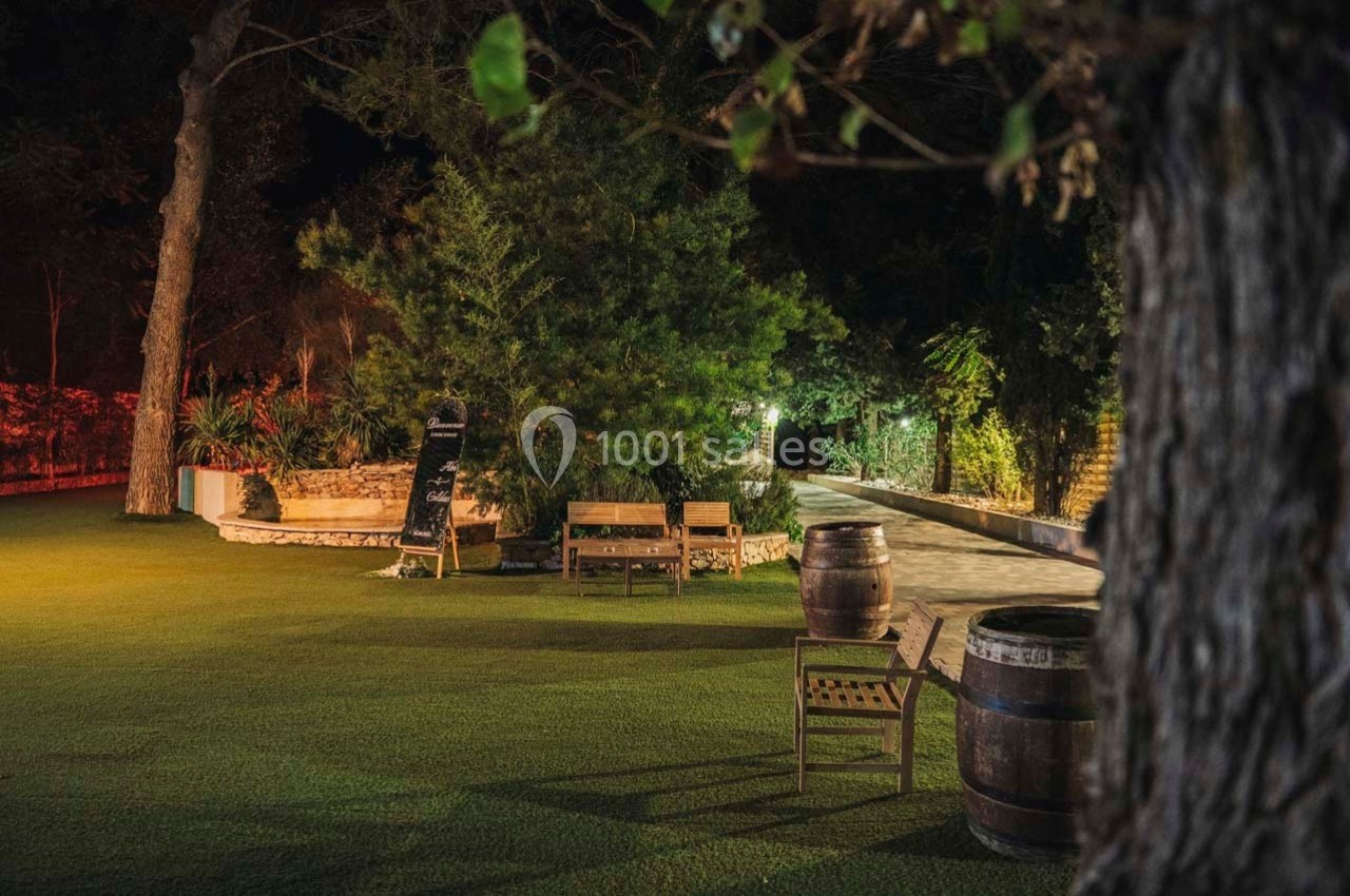 Espace extérieur nocturne avec bancs, tonneaux en bois, végétation dense et éclairage chaleureux.