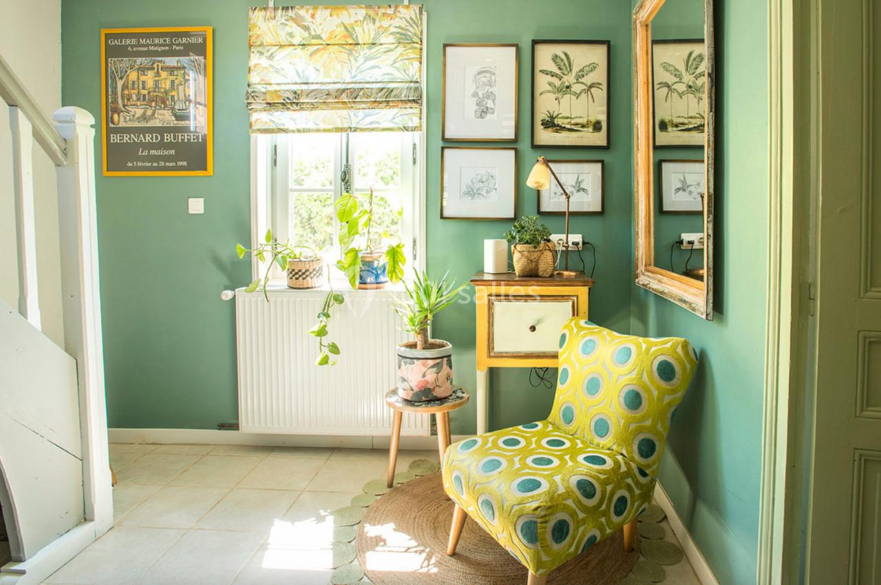 Coin de salon lumineux avec fauteuil jaune à motifs, plantes en pot, mur vert décoré de cadres et fenêtre avec store imprimé.
