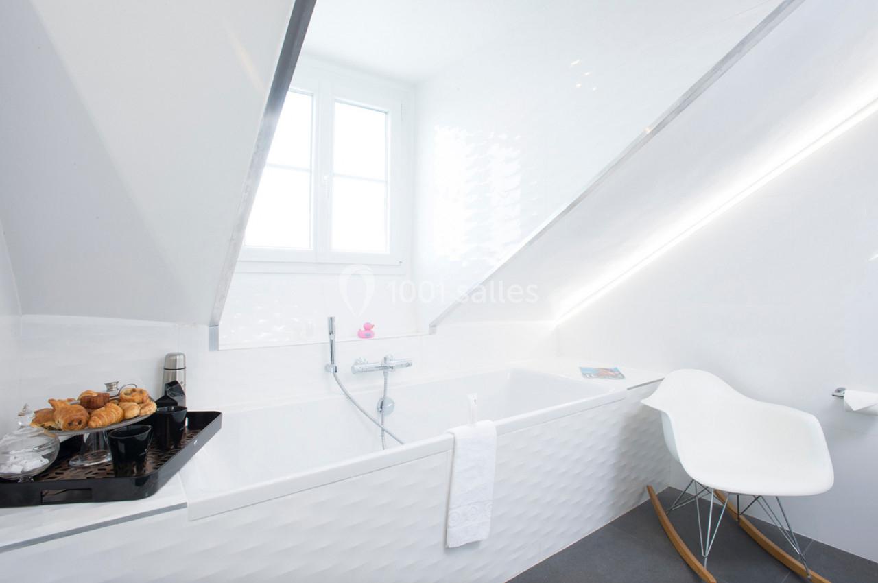 Salle de bain lumineuse sous combles avec baignoire, fenêtre, chaise blanche et plateau de viennoiseries.