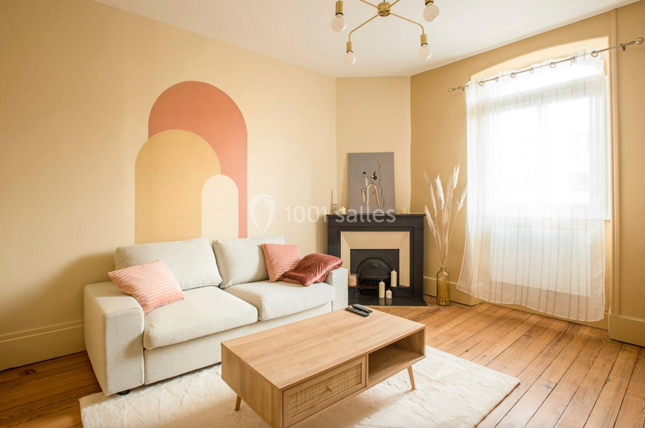 Salon lumineux avec canapé beige, coussins roses, table basse en bois, cheminée décorative et mur peint en arches colorées.