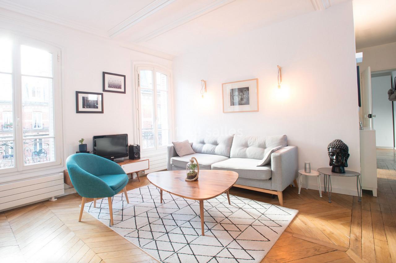 Salon lumineux avec parquet en bois, canapé gris, fauteuil bleu, table basse en bois et décoration murale minimaliste.