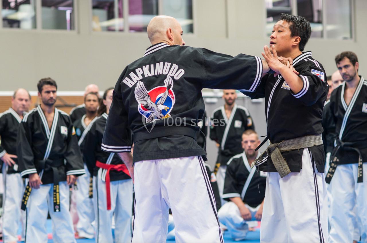 Deux hommes en tenue de hapkido s'entraînent dans un dojo, entourés d'autres pratiquants attentifs.