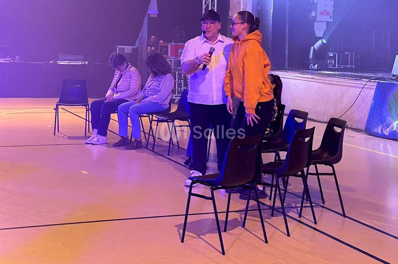 Un homme avec un micro interagit avec une femme debout, tandis que trois personnes sont assises sur des chaises en arrière…