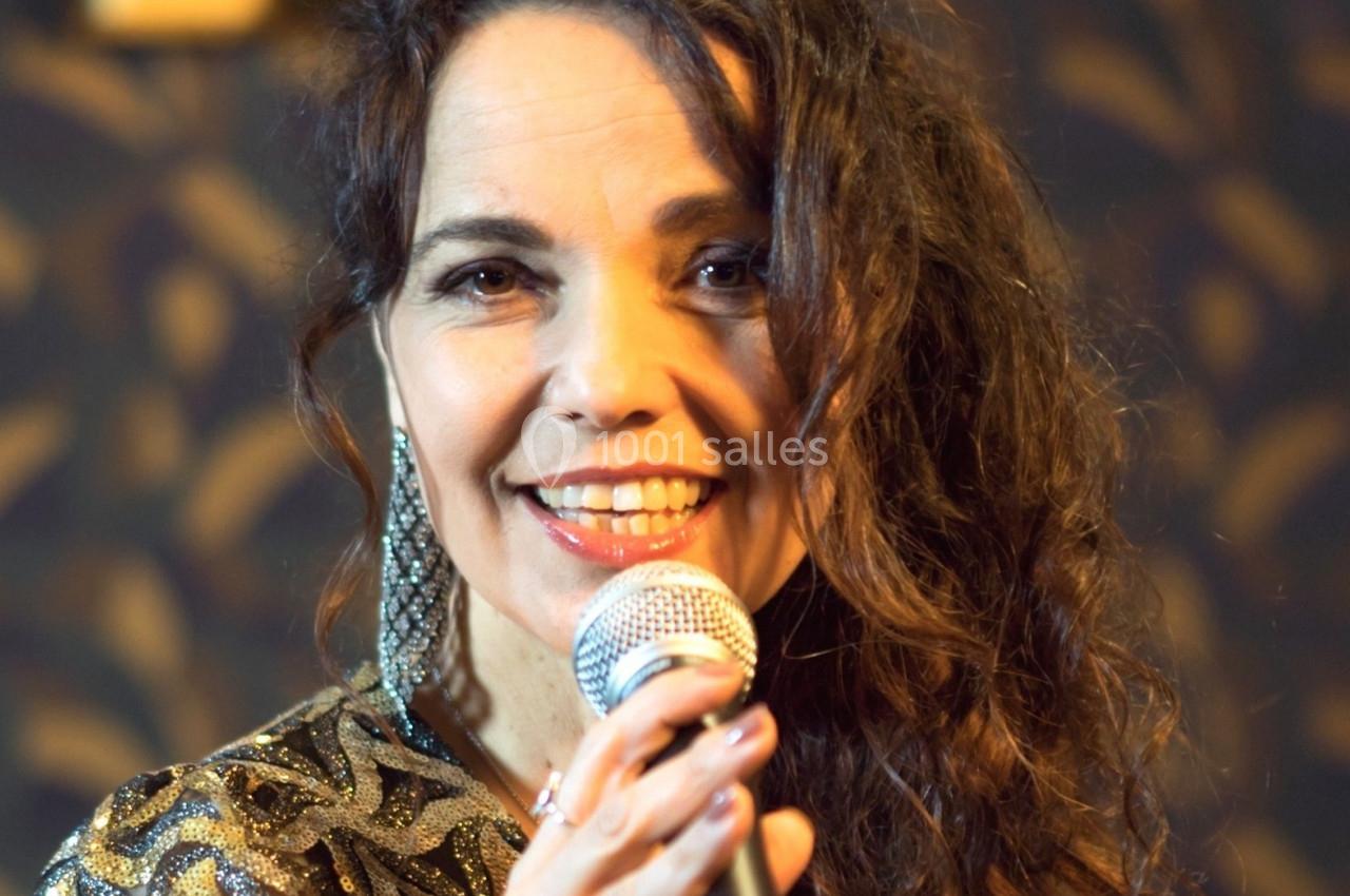 Femme souriante tenant un microphone, vêtue d'une tenue brillante, sur un fond sombre à motifs.