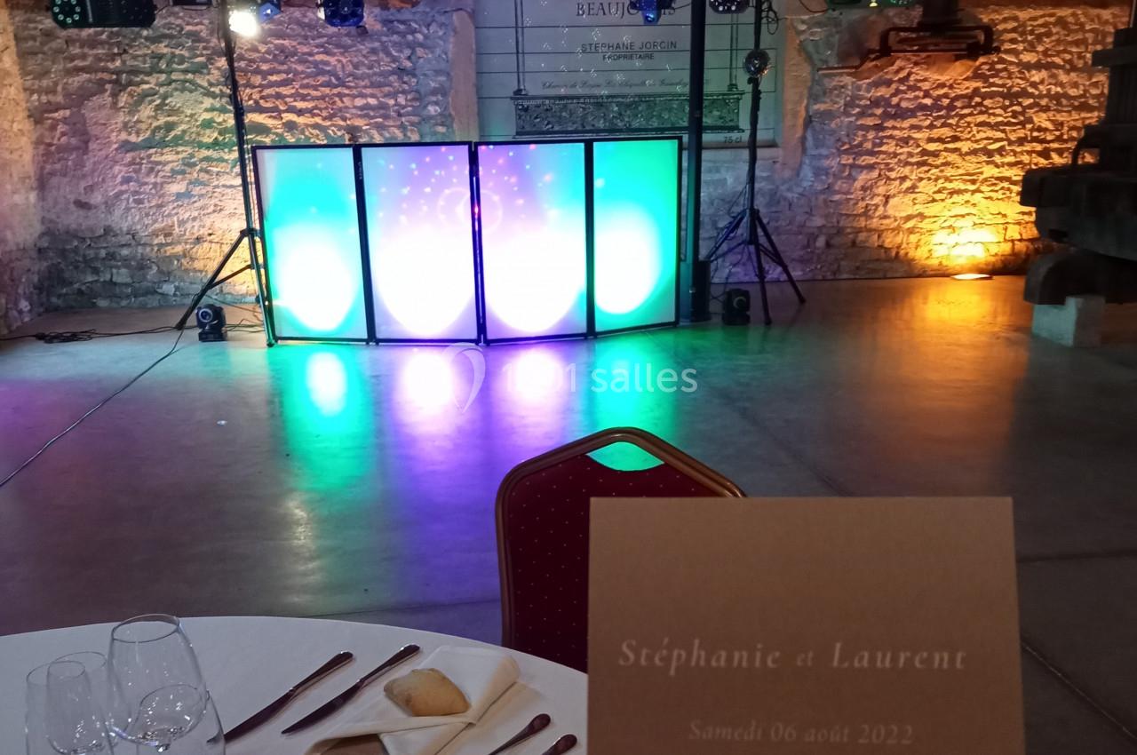Salle de réception éclairée avec une piste de danse colorée, une table dressée et un carton mentionnant des prénoms et une…