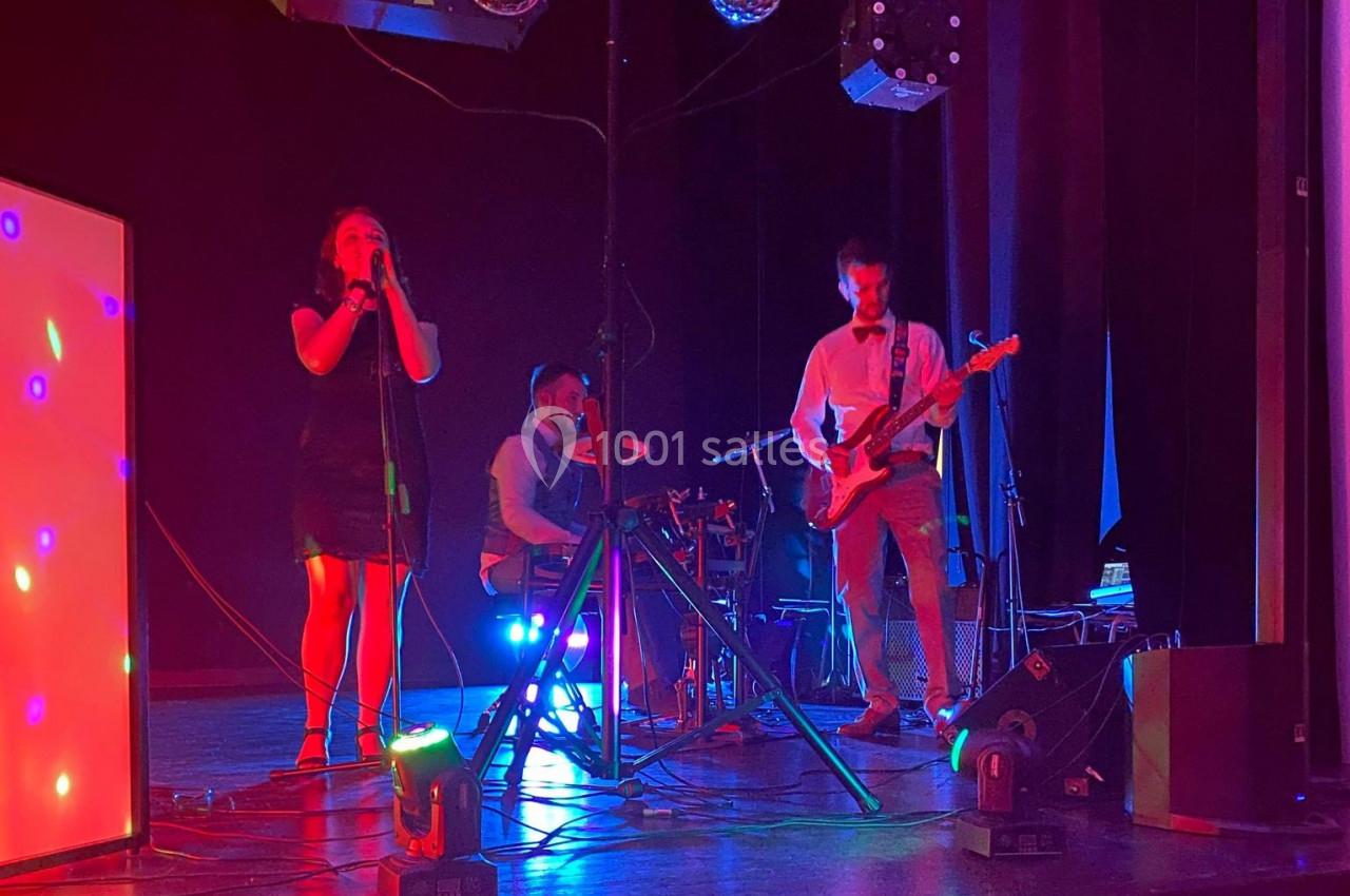 Un groupe de musiciens joue sur scène avec éclairages colorés, comprenant une chanteuse, un batteur et un guitariste.