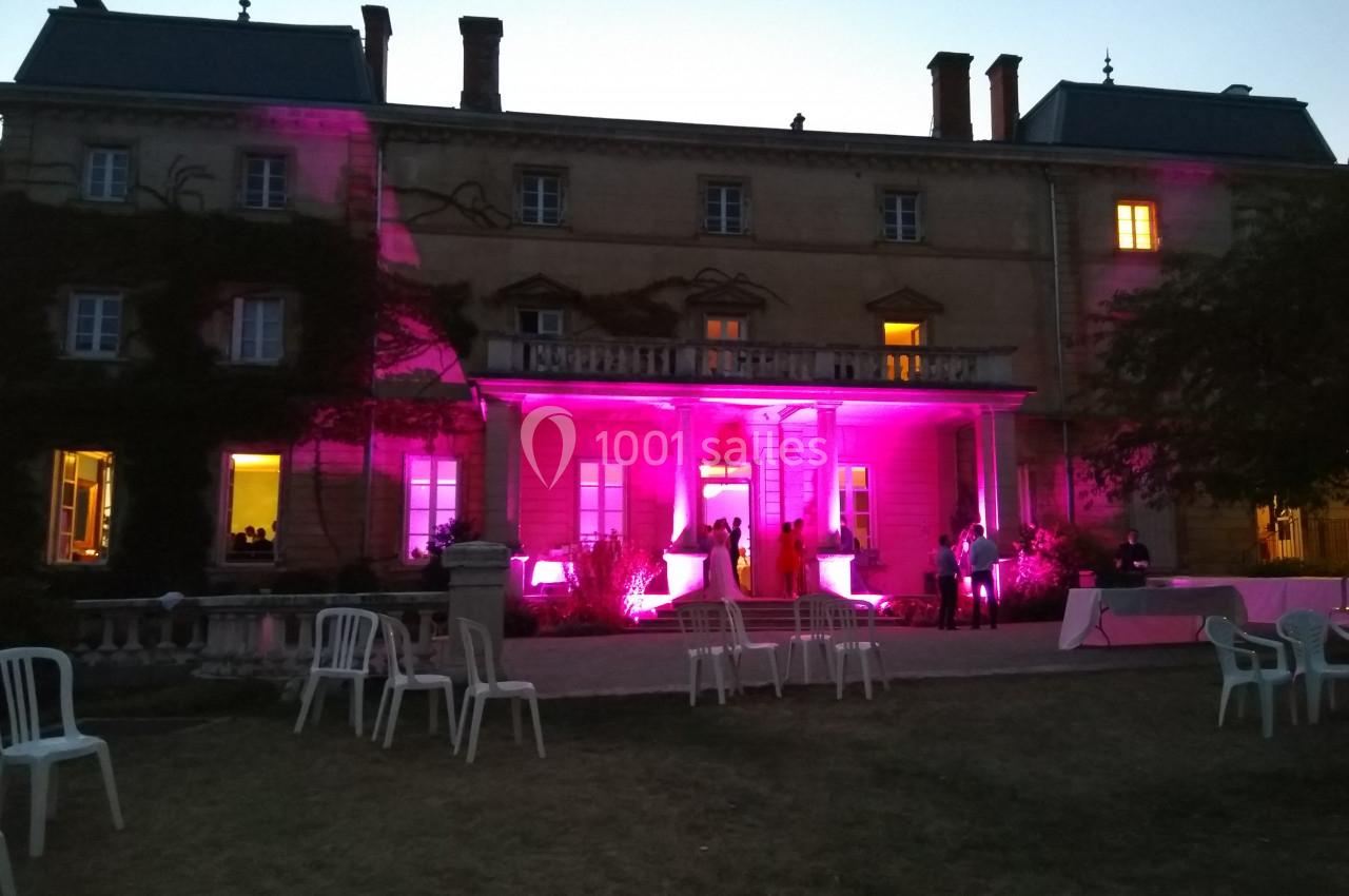 Façade d'un bâtiment éclairé par des lumières roses, avec des chaises blanches et des personnes dans le jardin au crépuscule.
