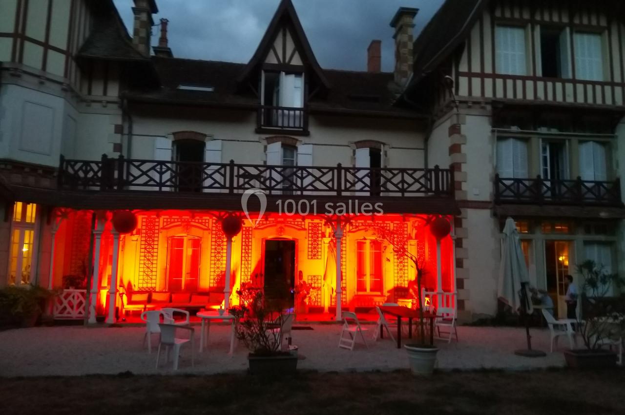 Façade d'une grande maison à colombages éclairée par des lumières rouges, avec mobilier de jardin au premier plan.