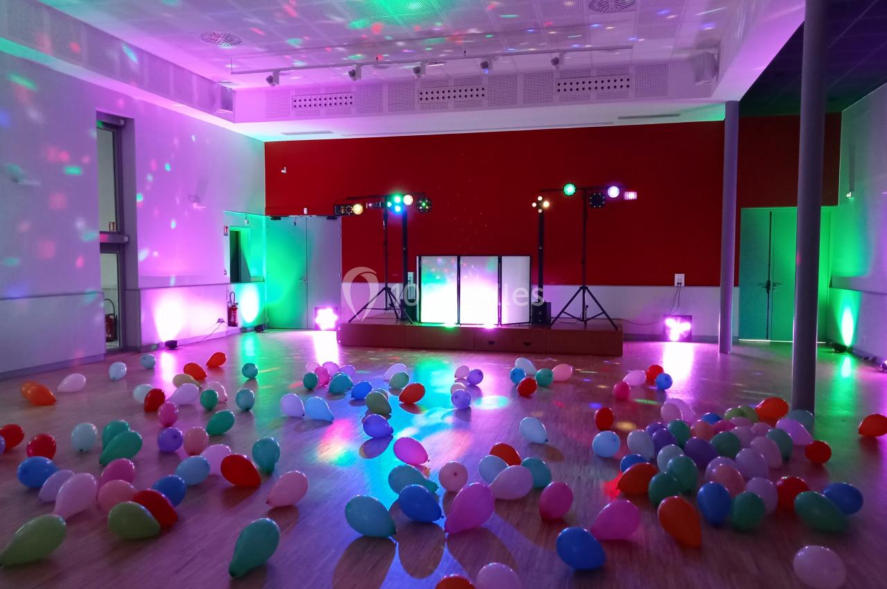 Salle de fête décorée avec des ballons colorés au sol et des lumières d'ambiance sur une scène équipée.