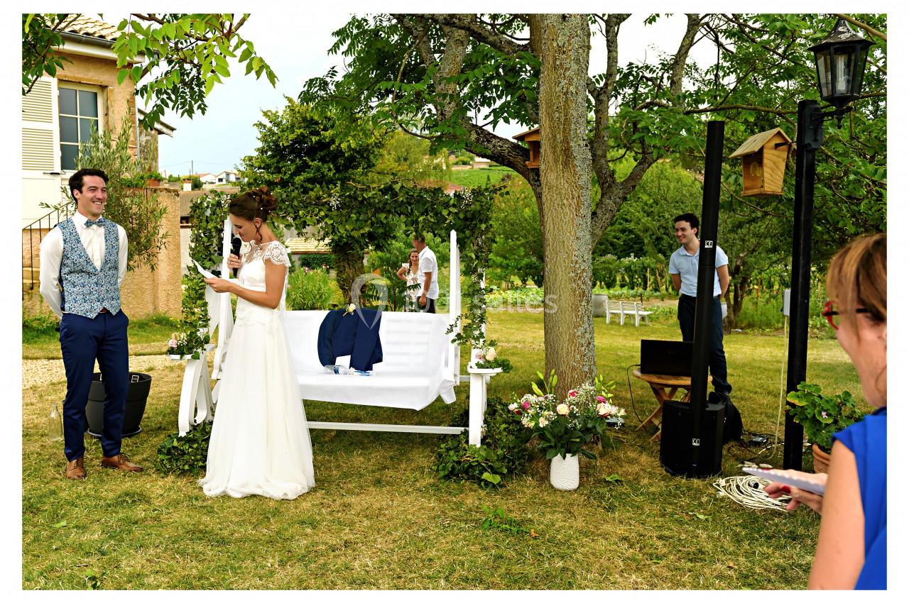 Une mariée lit un discours devant un marié souriant dans un jardin décoré pour une cérémonie en plein air.