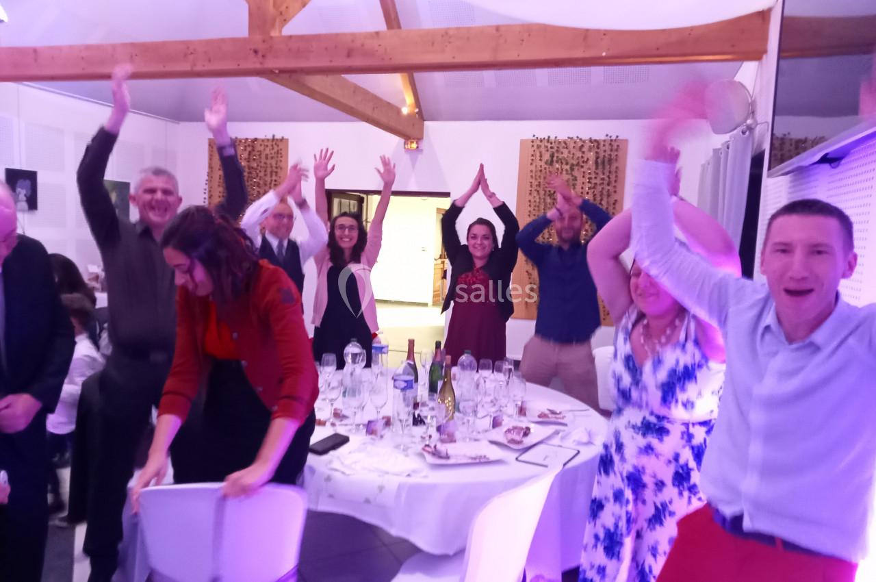 Des personnes souriantes debout autour de tables décorées, levant les bras dans une ambiance festive en intérieur.